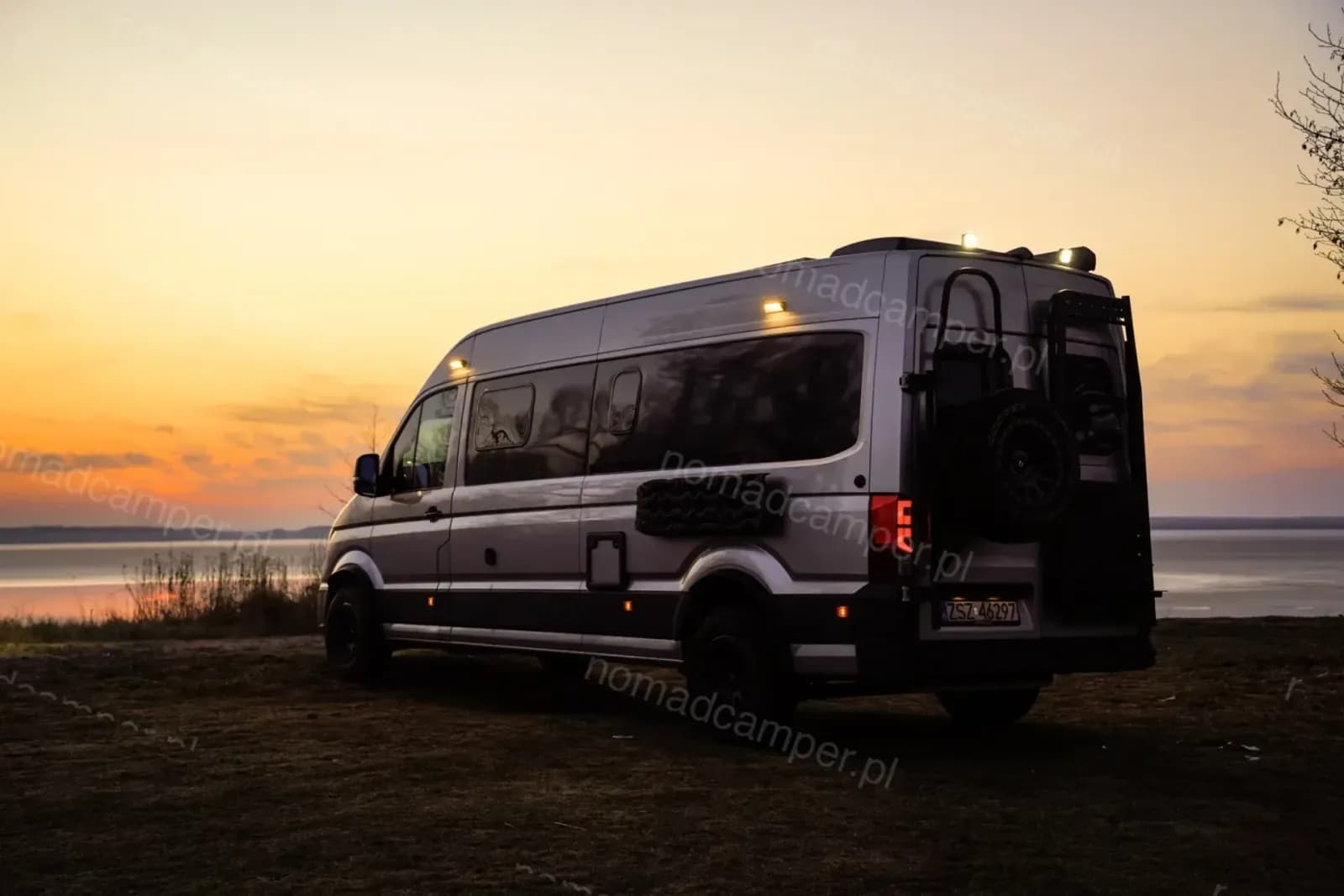 Nomad Camper MAN TGE nad jeziorem o zachodzie słońca, z włączonymi światłami dachowymi